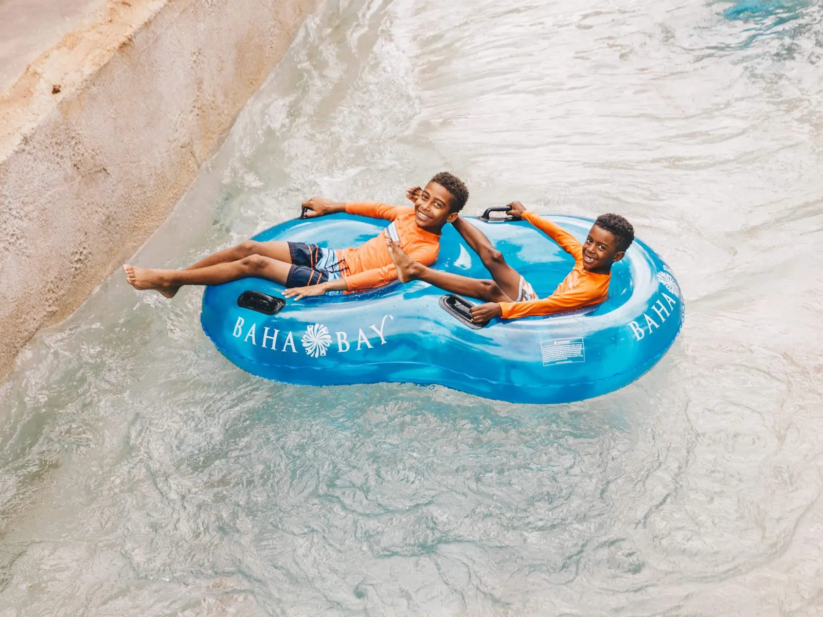 2 children in orange tops on a blue double tube marked Baha Bay, in the current of the water park’s river, seen from above.