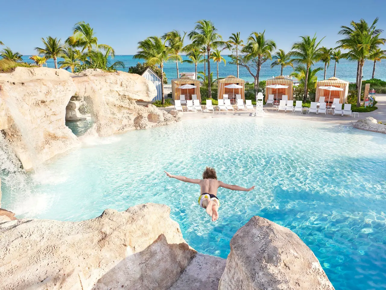 A boy dives from a rock into the Dean’s Blue Hole pool; opposite, a row of yellow-striped cabanas, palms and the sea.