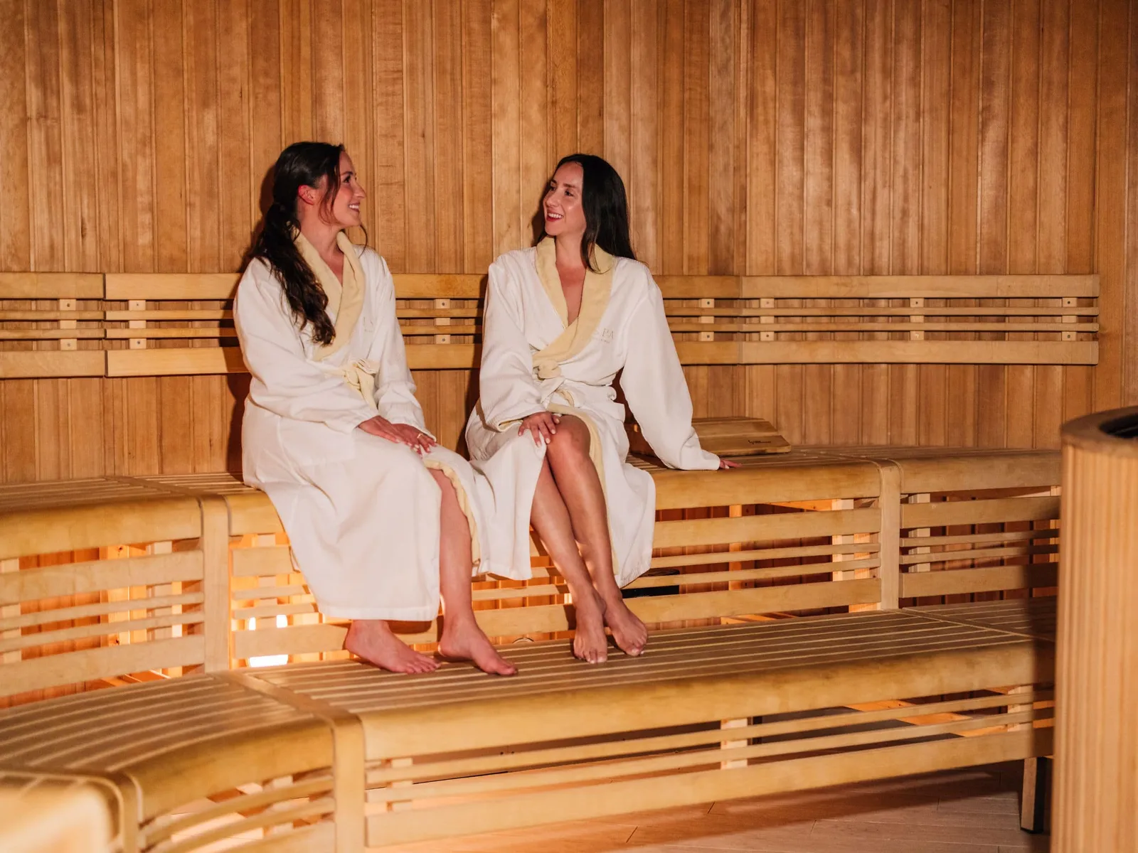 2 women in white robes sitting on the pale wooden benches of a sauna at the ESPA spa.