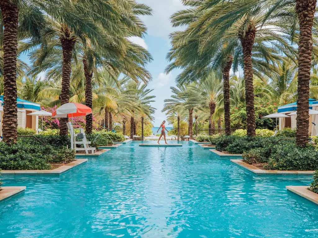 A long, narrow pool between 2 rows of palms, with striped umbrellas at the sides and, at the far end, one figure on the edge in front of the sea.