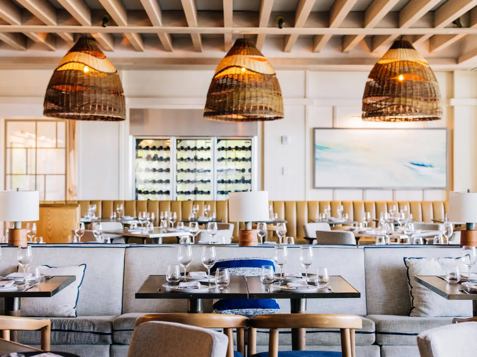 The dining room of Marcus at Baha Mar Fish + Chop House: laid tables, pale banquettes, 3 large wicker lamps and a wall of wine bottles.
