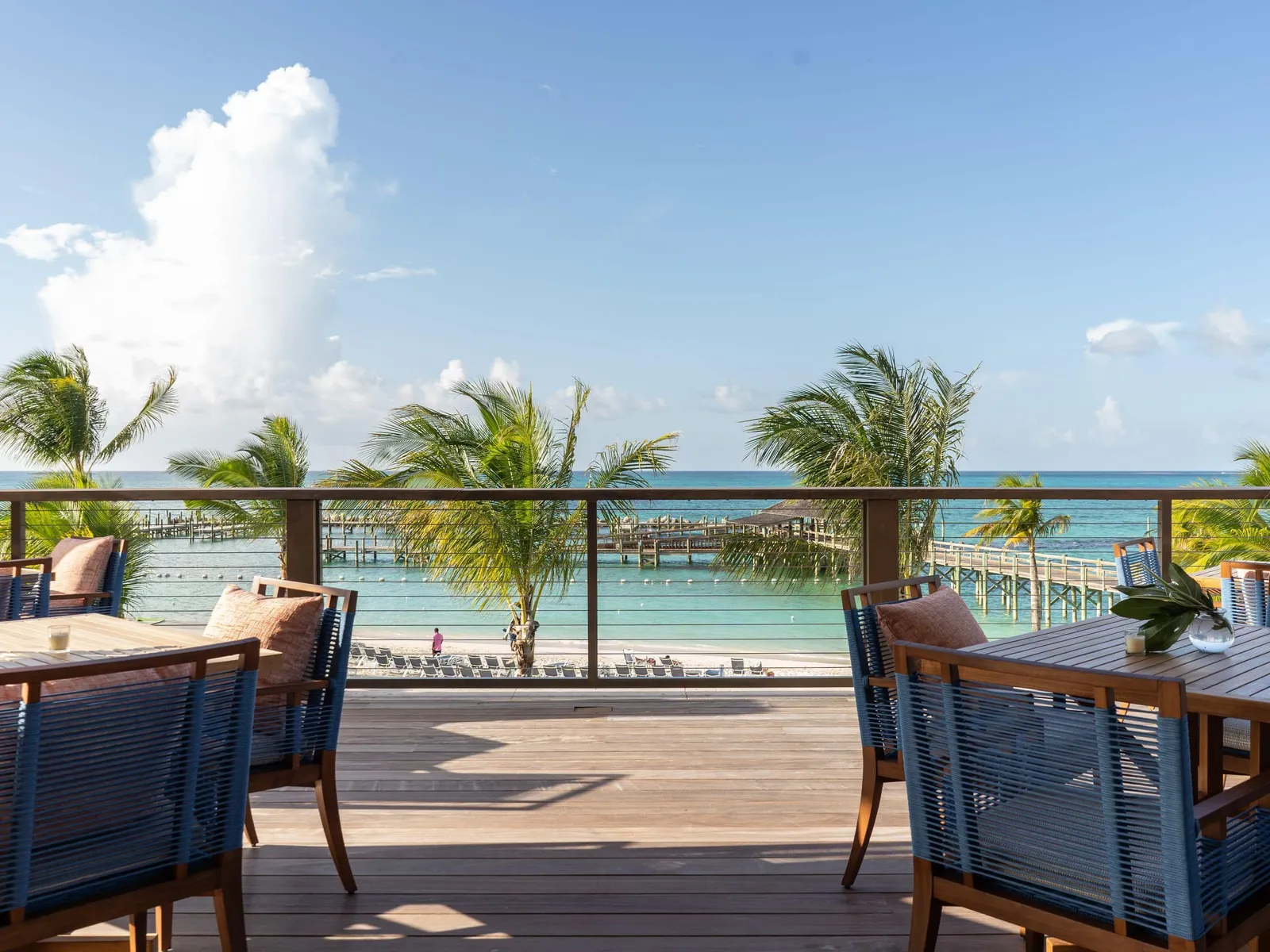 The wooden terrace at Marcus: tables with woven blue chairs and, beyond the railing, palms, the beach and the pier over the sea.