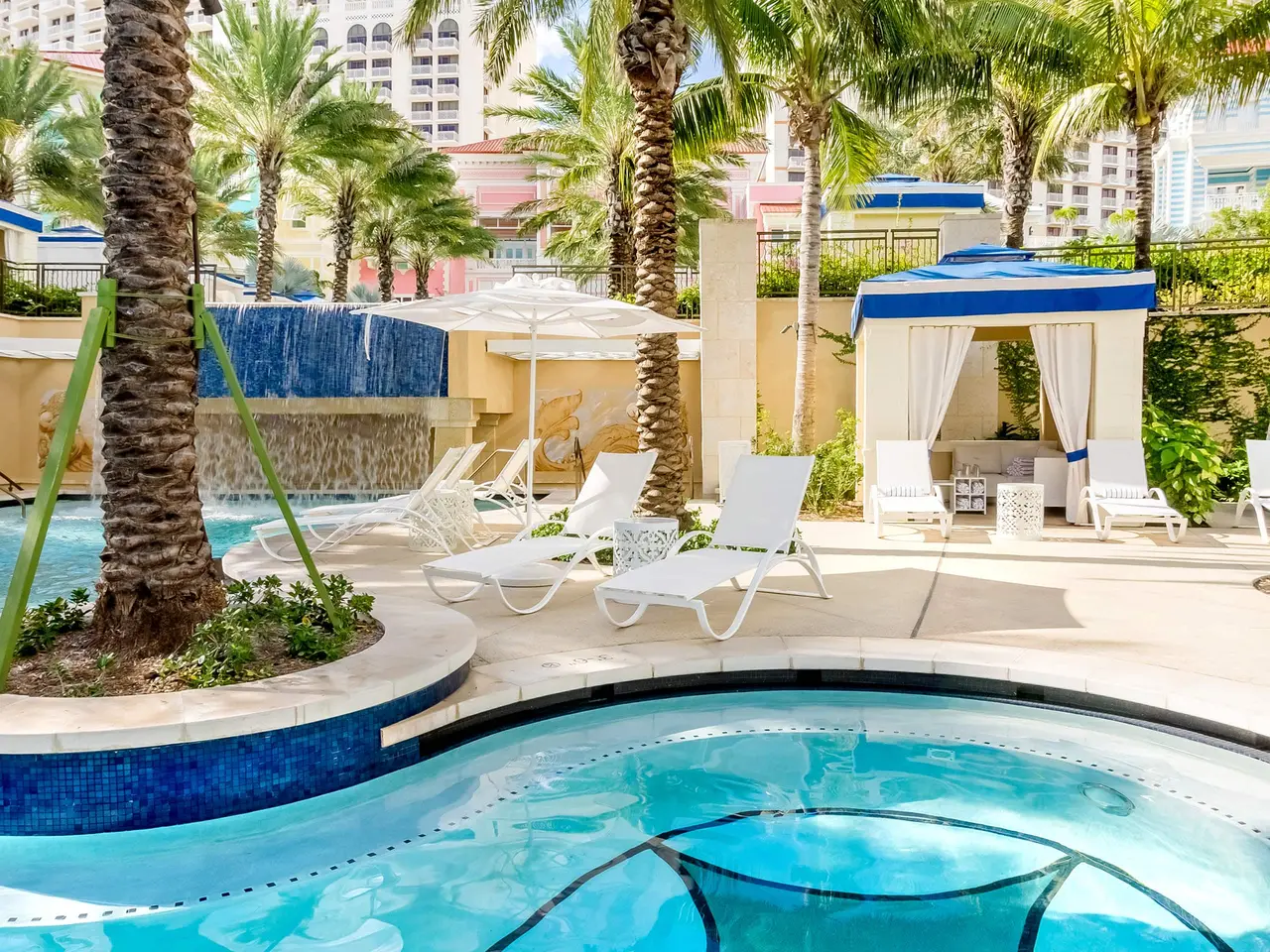 A corner of the Fortune pool: a round basin in blue mosaic, white loungers, a cabana with white and blue curtains and a waterfall among the palms.
