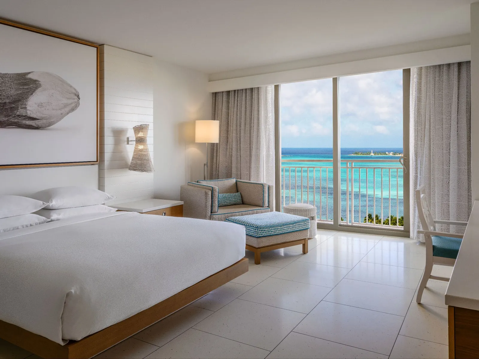 An ocean-view king room: the bed, the armchair with a pale-blue footstool and the railed glass door onto the turquoise sea.