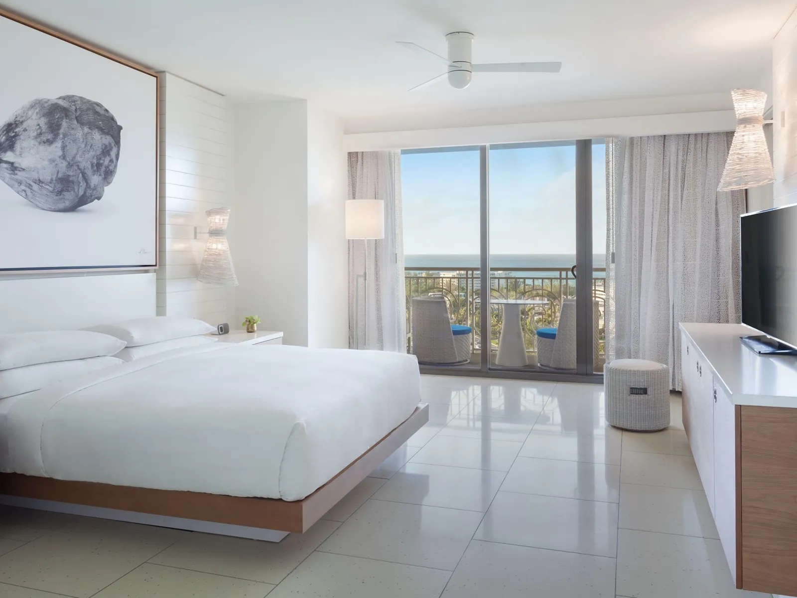 A king room with a balcony: the bed in the foreground, the television cabinet and, outside, a small table with 2 blue chairs facing the sea.