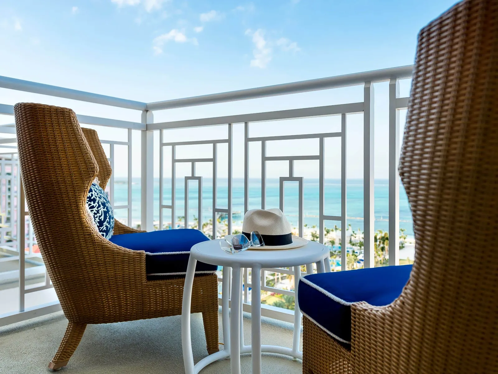 The balcony of an East Tower room: 2 high-backed wicker armchairs with blue cushions, a small table with a straw hat and the sea beyond the white railing.
