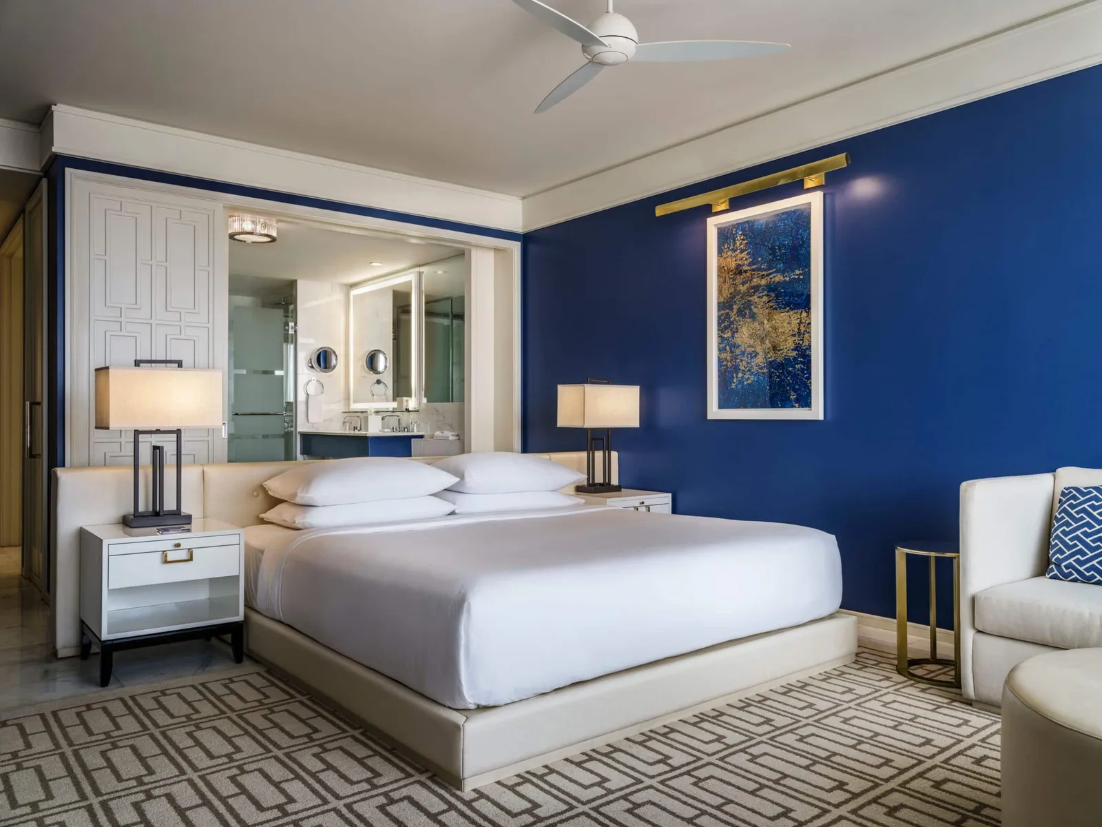 A Deluxe king room in the East Tower: dark-blue walls, the white bed on a geometric rug and, behind it, the open bathroom with its lit mirror.