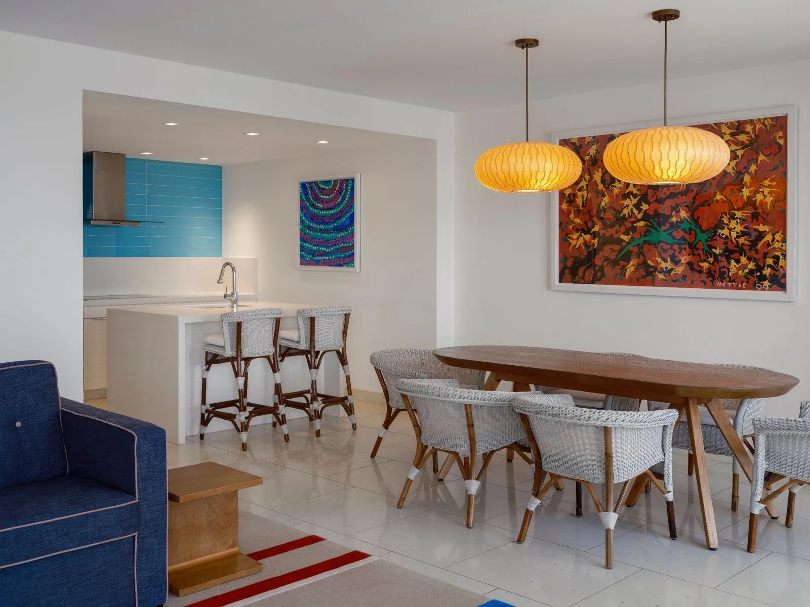 The dining area of a Deluxe residence: an oval wooden table with 6 wicker chairs under 2 yellow lamps, a large red painting and the turquoise kitchen.