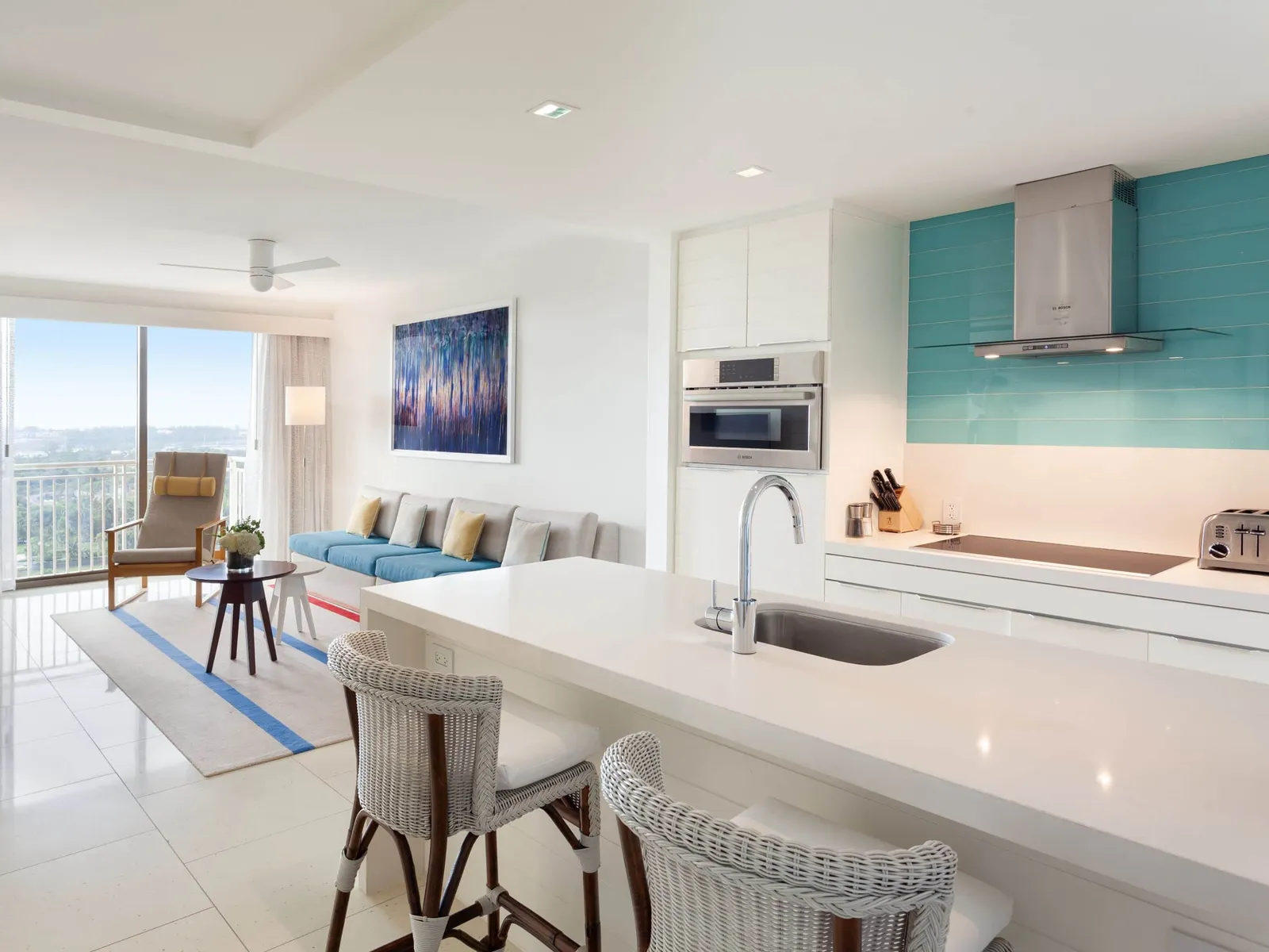 The kitchen of a one-bedroom residence in the West Tower: the white island with its sink and 2 wicker stools, the turquoise wall and the living room with its balcony.