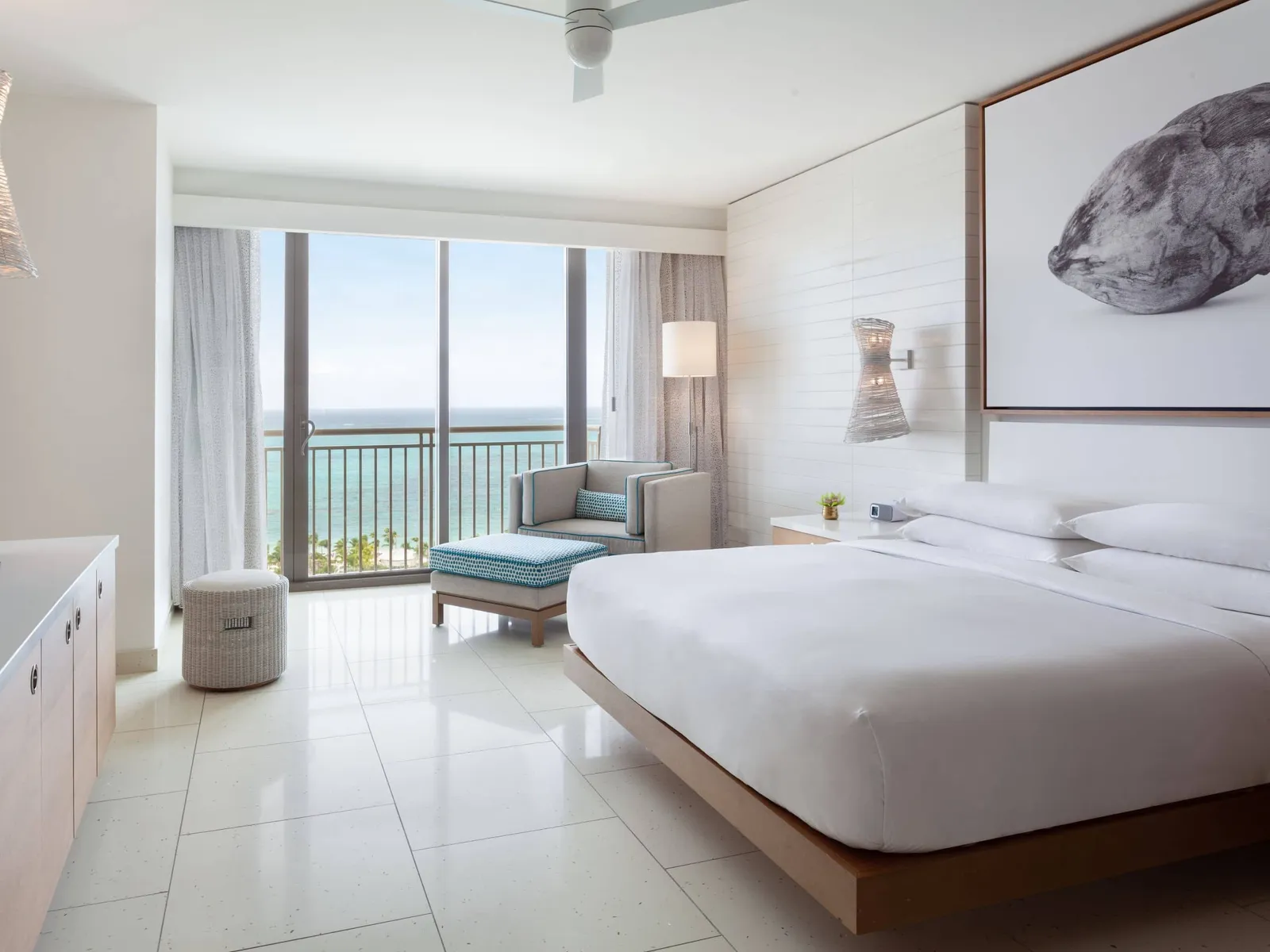The bedroom of an ocean-view Deluxe residence: the king bed under the photograph of a shell, the pale-blue chaise and the sea beyond the glass door.