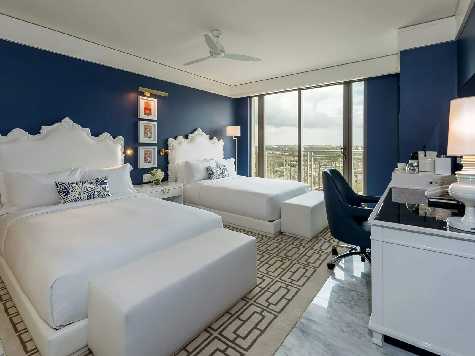 2 queen beds with white benches at their feet, 3 small pictures on the blue wall and the window over the island’s interior.
