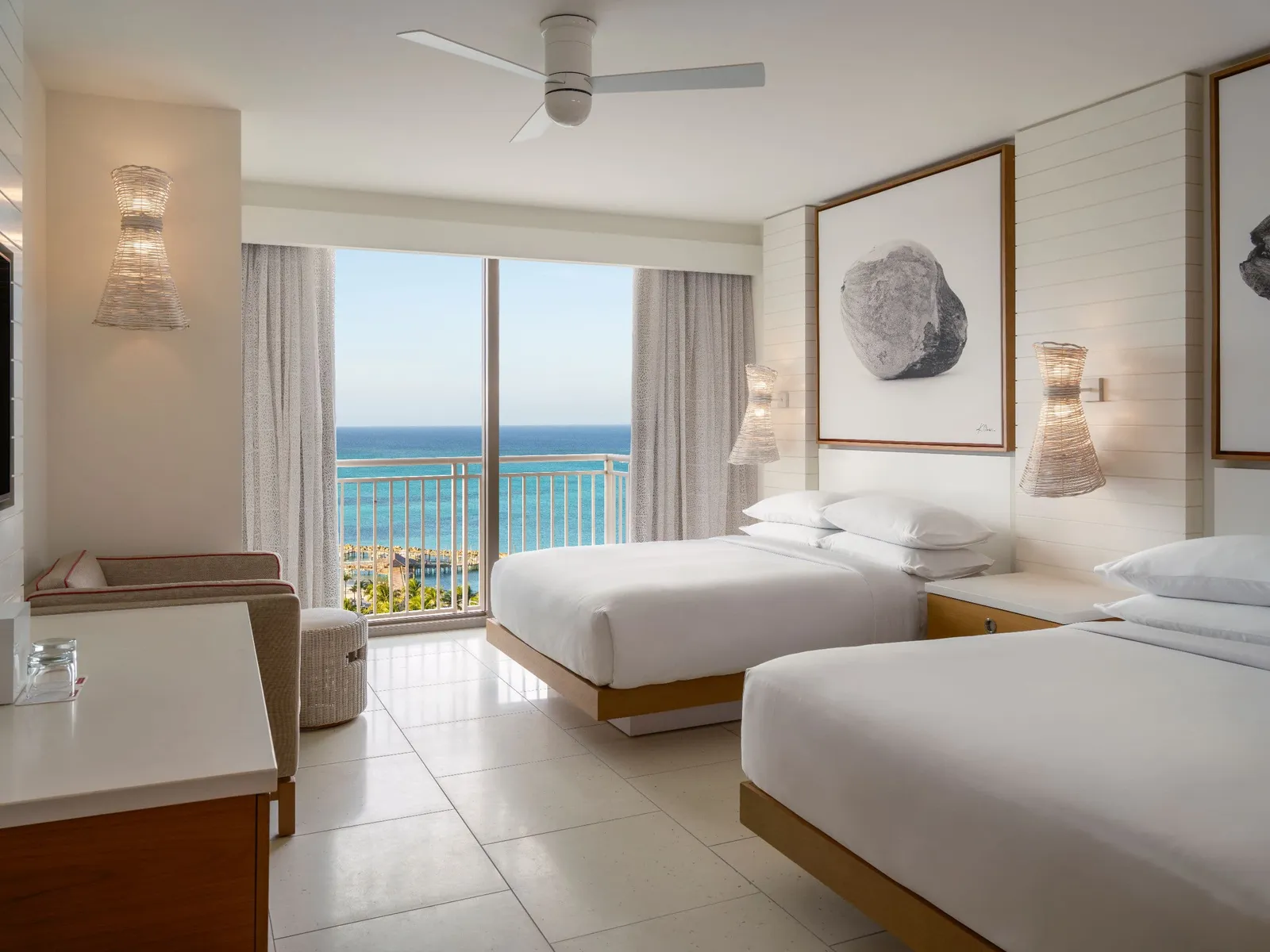 2 queen beds with wicker wall lights at their sides and, beyond the glass door, the coast and the sea off Cable Beach.