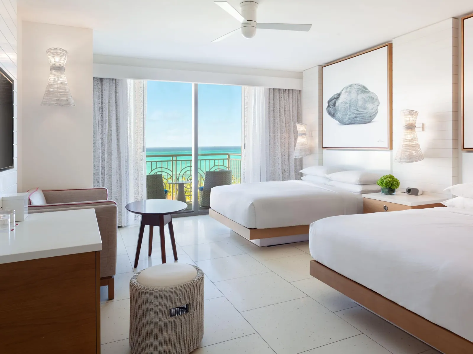 A room with 2 queen beds and a small round table, the glass door open onto the balcony and the turquoise sea.