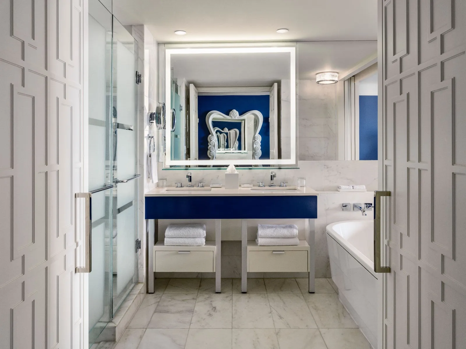 The bathroom of an East Tower Deluxe room: the blue double vanity under a lit mirror, the glass shower on the left and the bathtub on the right.