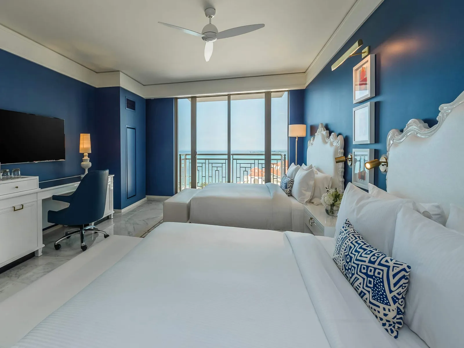 2 queen beds with scalloped white headboards against blue walls, the desk with its blue chair and the windows over the sea.