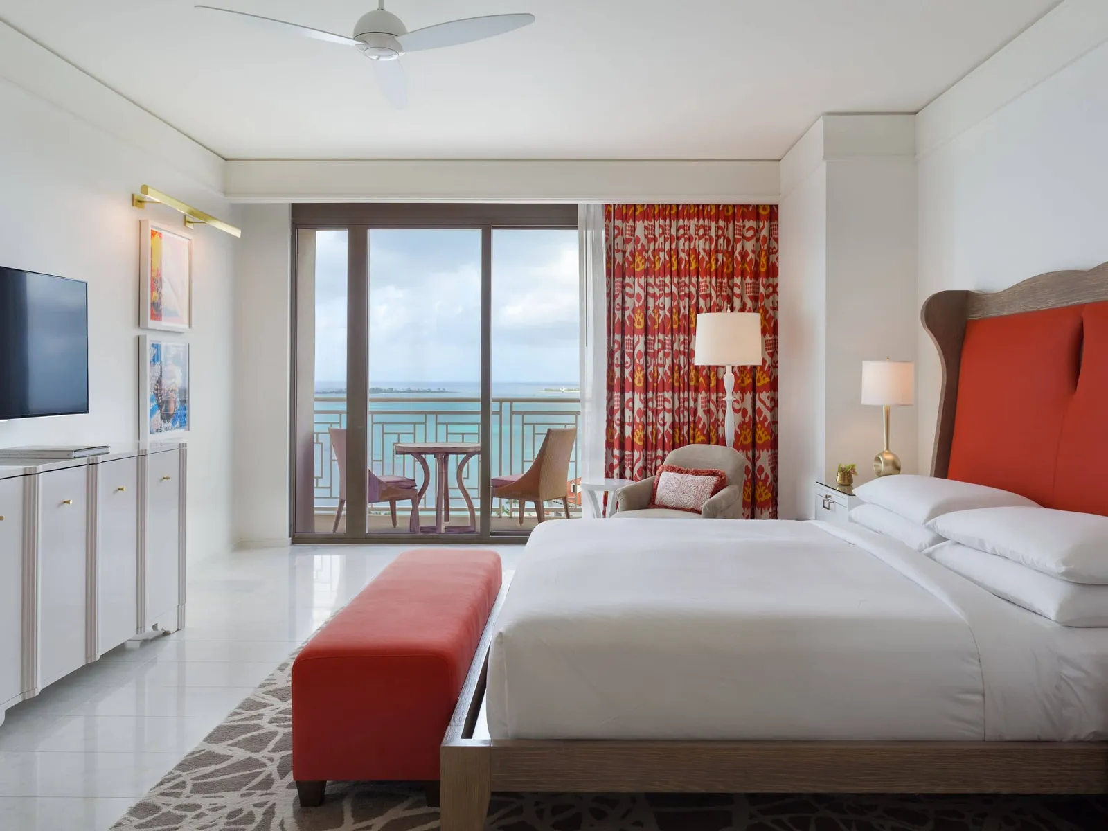 The bedroom of an ocean-view Reserve residence: the bed with its red headboard, the red bench at its foot and the balcony with 2 chairs facing the sea.