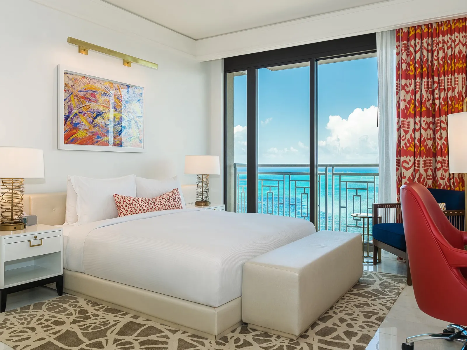 A bedroom of the Vista residence: the white bed with a red-patterned cushion, the red desk chair and the balcony over the turquoise sea.