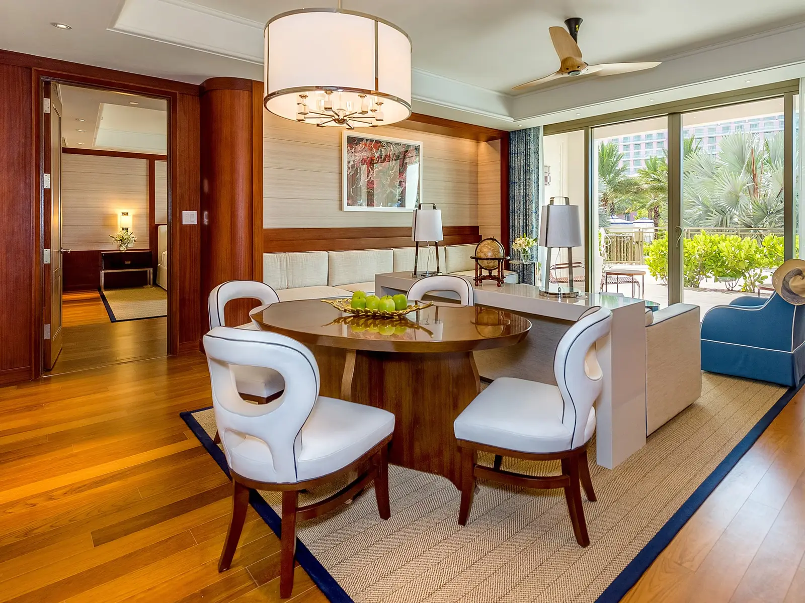 The living room of a poolside villa: wooden floor, the round table with white chairs, the pale sofa and the glass door to the terrace among the palms.