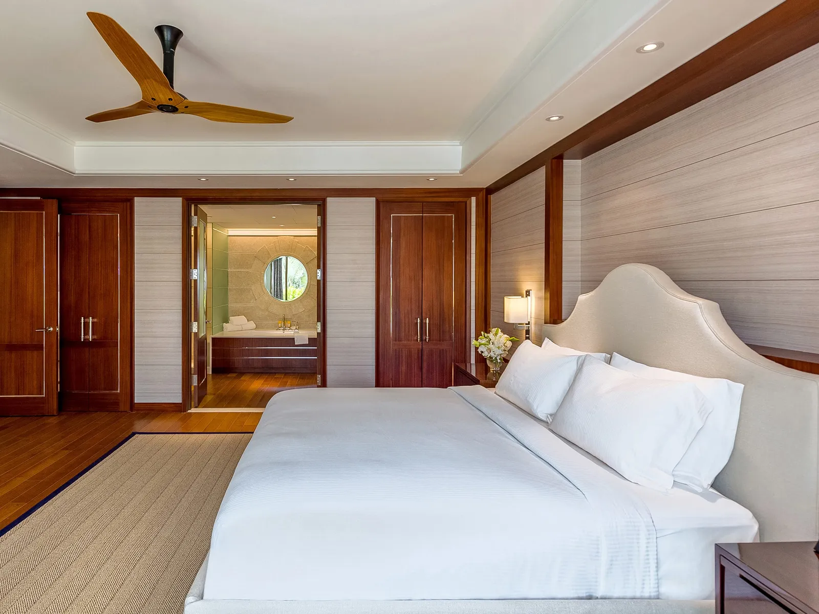 The bedroom of a poolside villa: the king bed with its pale headboard, wood-panelled walls, the wooden-bladed ceiling fan and, beyond, the bathroom.