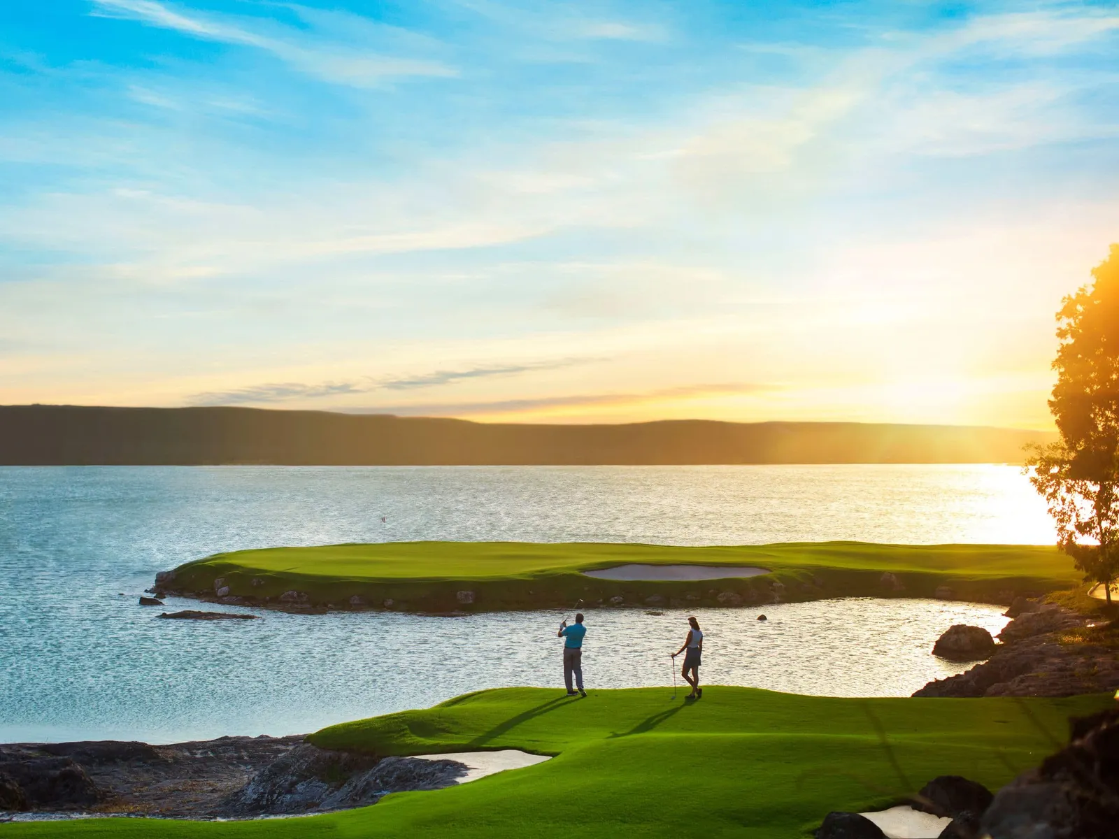 2 golfers at sunset on a Royal Blue green surrounded by water, the sun low behind the trees.