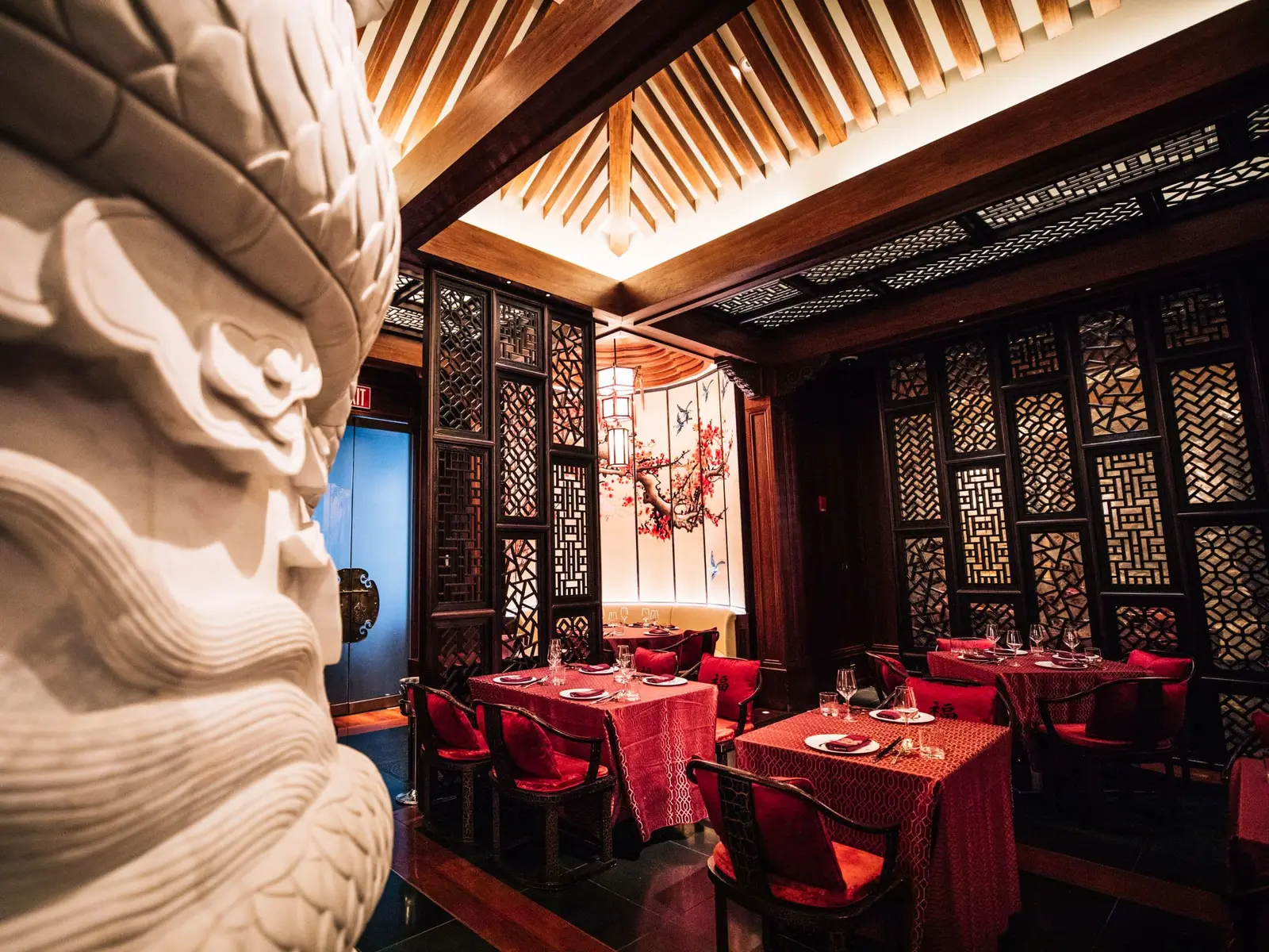 Shuang Ba’s dining room: tables with red cloths between carved wooden screens, under a slatted ceiling, with a white stone sculpture in the foreground.