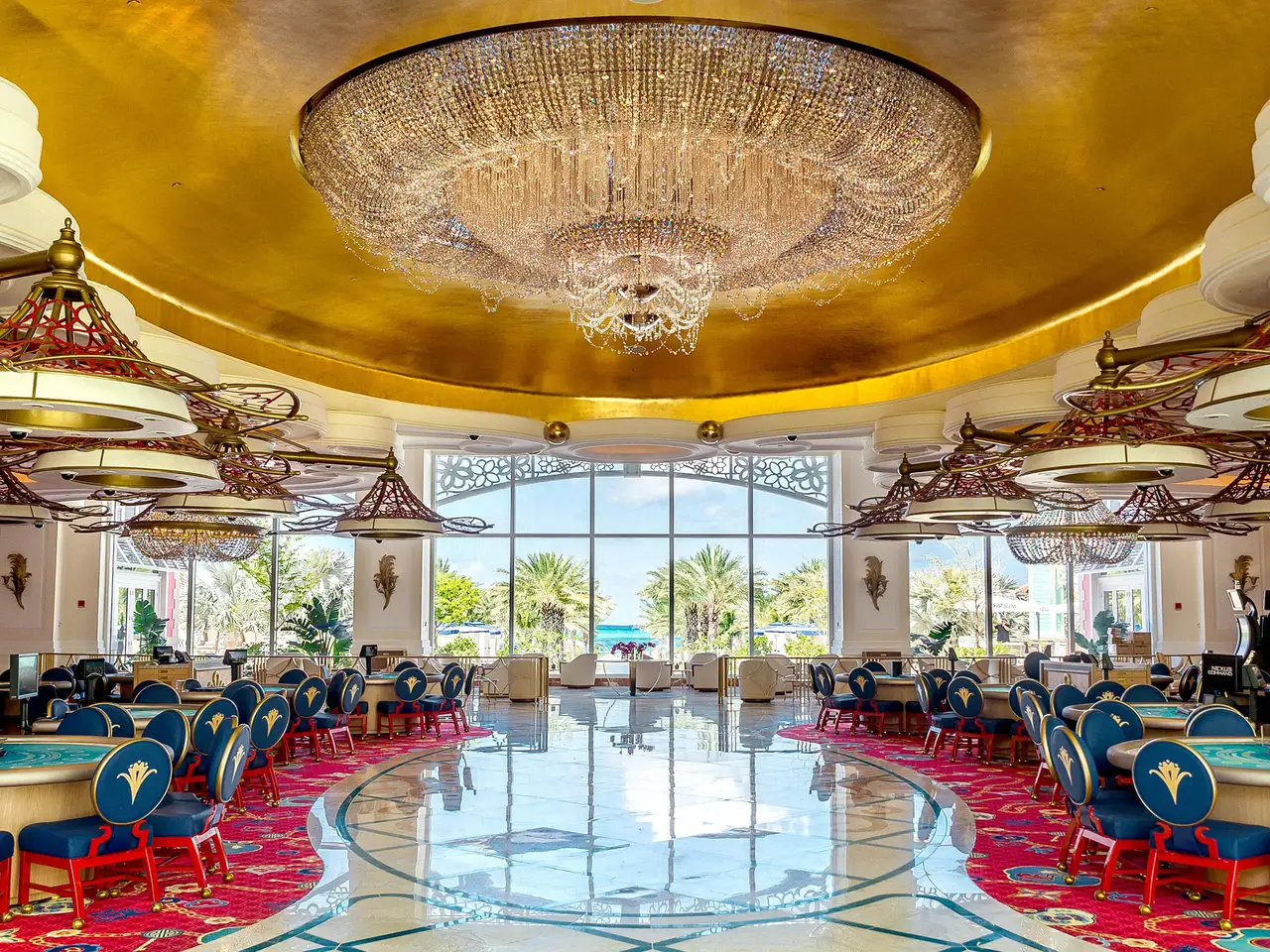 The casino floor, empty: a crystal chandelier under a gilded ceiling, gaming tables with blue chairs, a patterned red carpet and tall windows onto the gardens.
