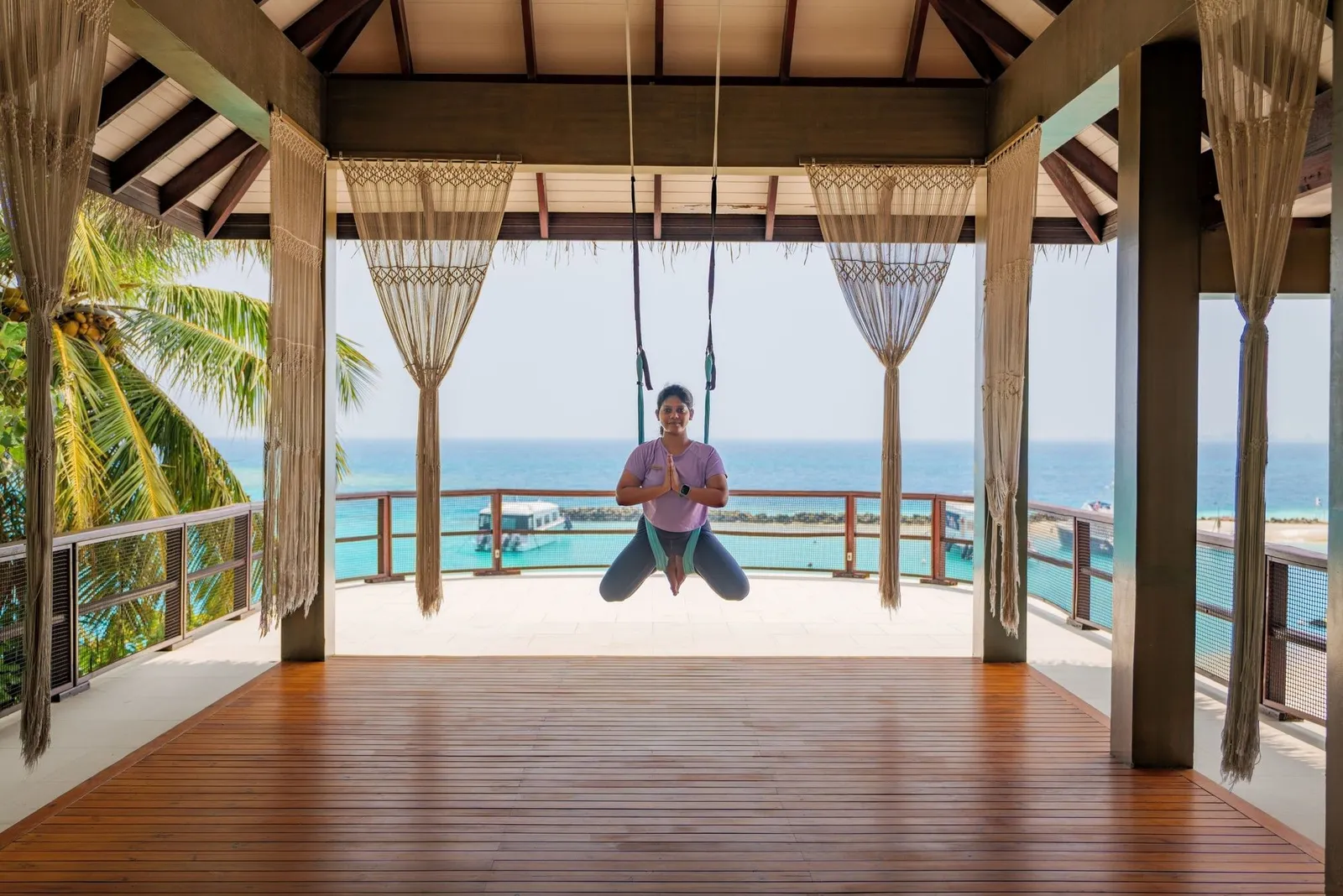 Aerial yoga in the pavilion facing the sea: a teacher suspended in a fabric hammock between the timber posts.