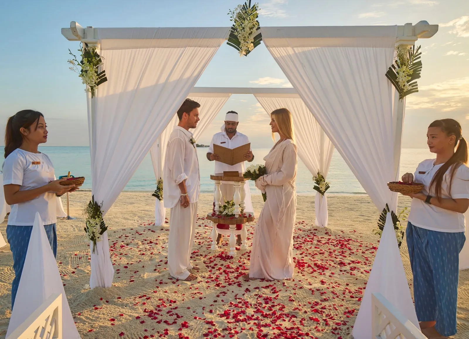 A ceremony on the beach: the couple under the white canopy, petals on the sand and 2 attendants holding trays.