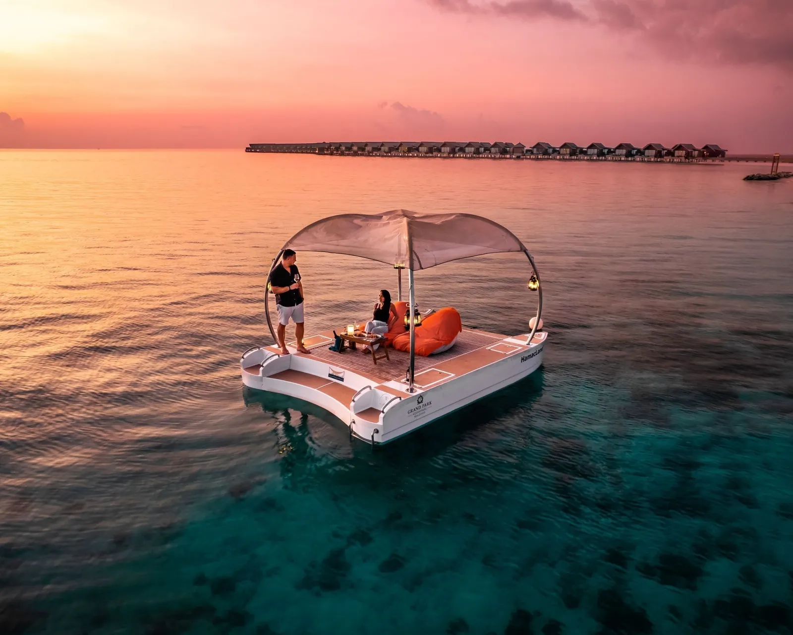 The floating deck at sunset, with its canopy, 2 guests and a waiter, and the water villas on the horizon.