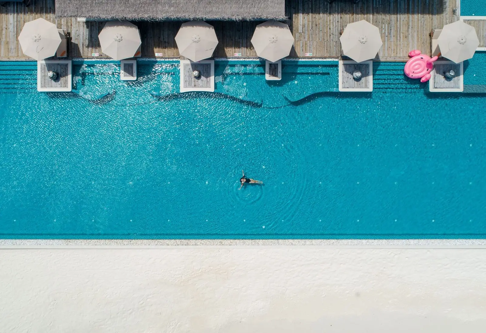 The infinity pool from above: a woman swimming in the blue water, the parasols along the timber edge and the white sand.