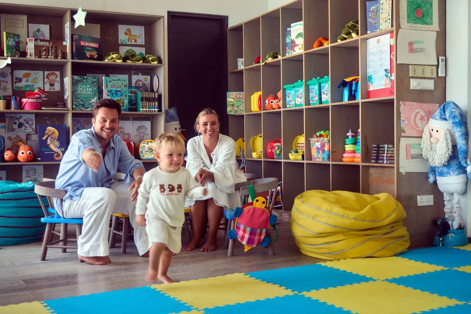 The kids’ club: a small child runs across the coloured mat in front of his parents, among shelves of toys and books.