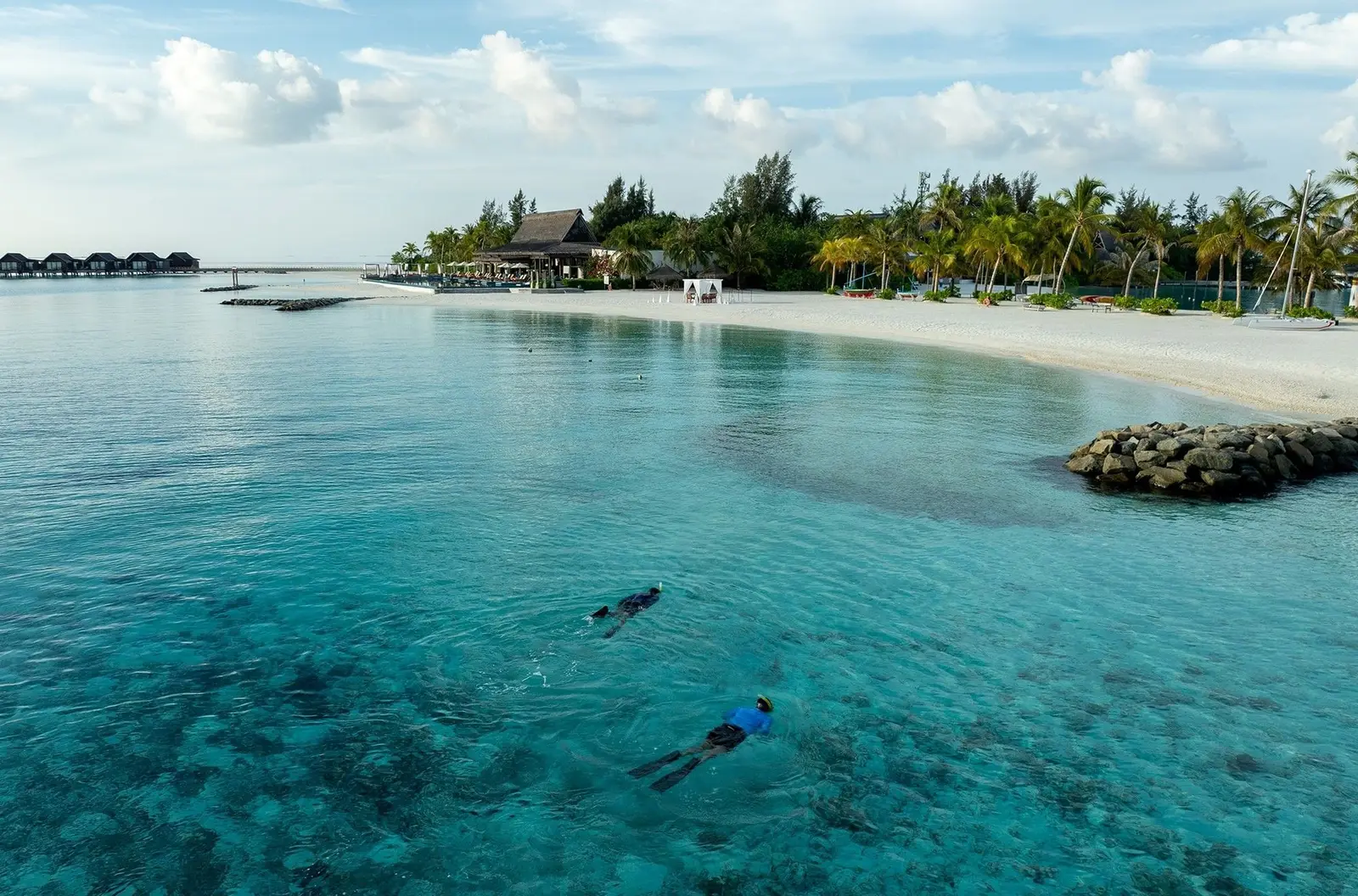 2 people snorkelling in the clear shallow water near the beach, with the water villas in the distance.