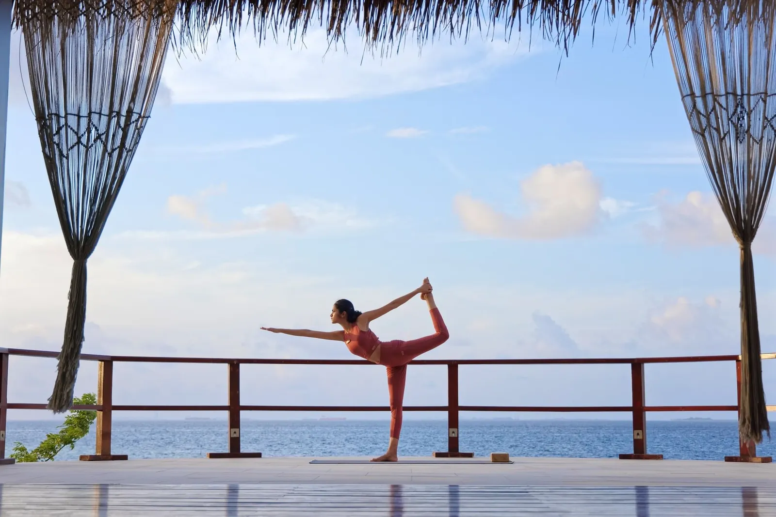 A woman holding a yoga pose on the timber deck of the pavilion facing the ocean, between 2 macramé curtains.
