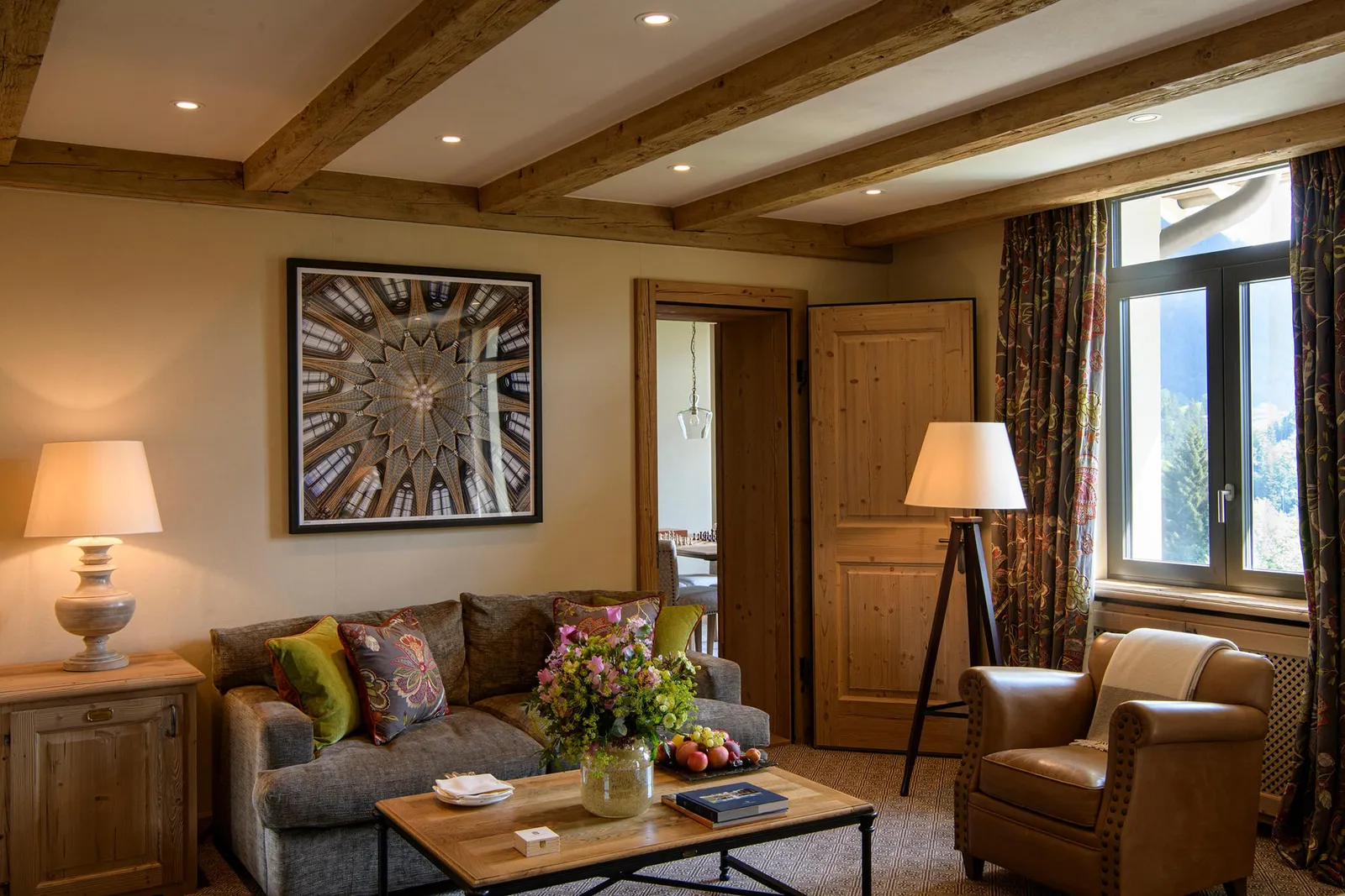 The living room of an Alpine Suite Mountain View: a grey sofa with coloured cushions, a leather armchair, a coffee table with flowers and fruit and a window onto trees