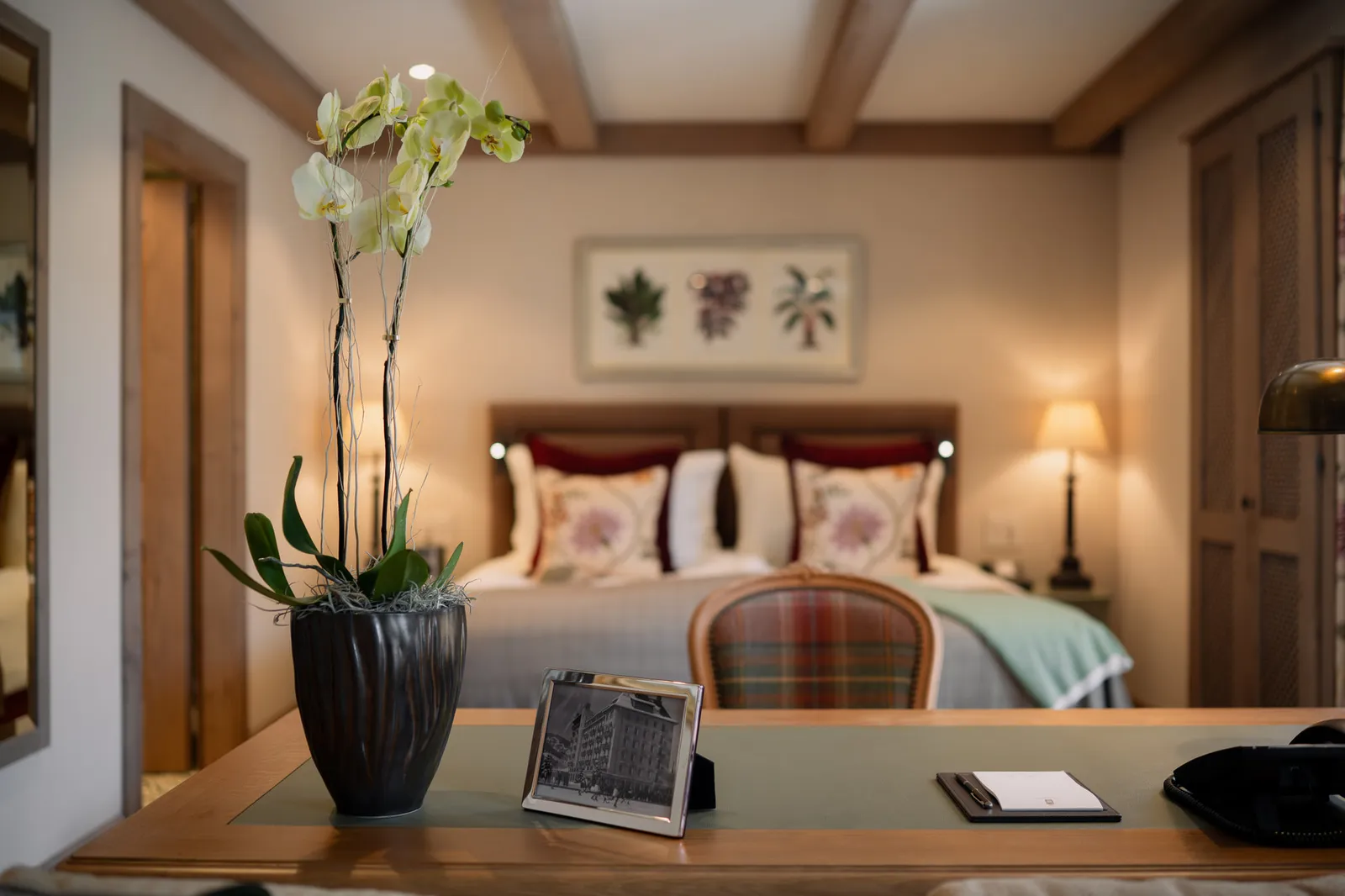 A Classic Junior Suite: a white orchid on the desk in the foreground and, behind it, the bed with floral cushions, a botanical print and exposed beams