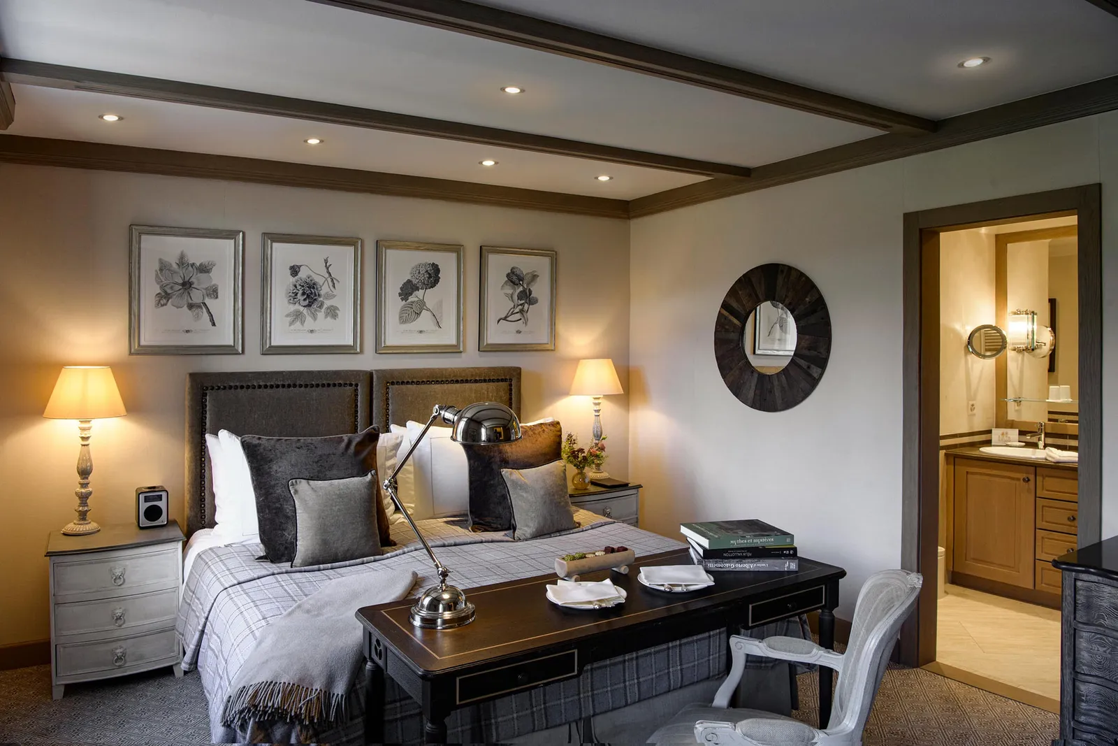 A Classic Room: a bed with a tartan throw, 4 black-and-white botanical prints, a round mirror, a desk with a brass lamp and the bathroom door open