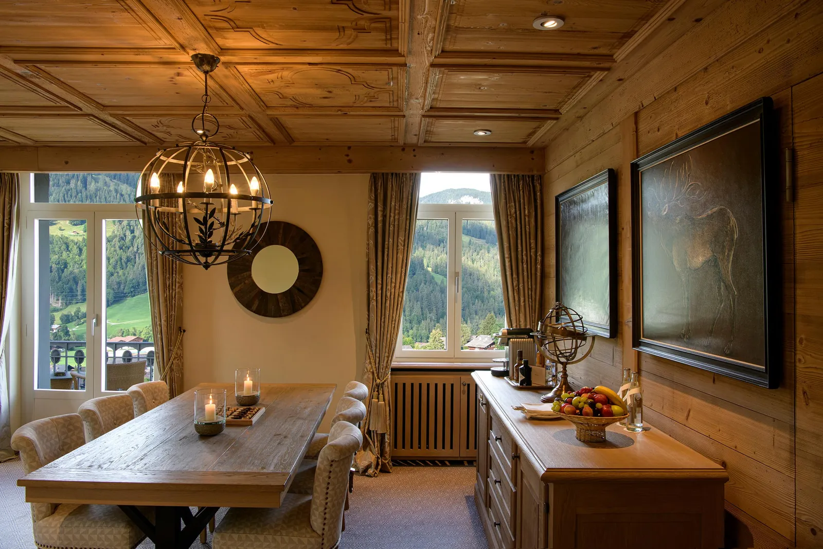 The dining room of a Corner Suite Mountain View: a wooden table under an iron chandelier, a carved coffered ceiling and 2 windows onto wooded slopes