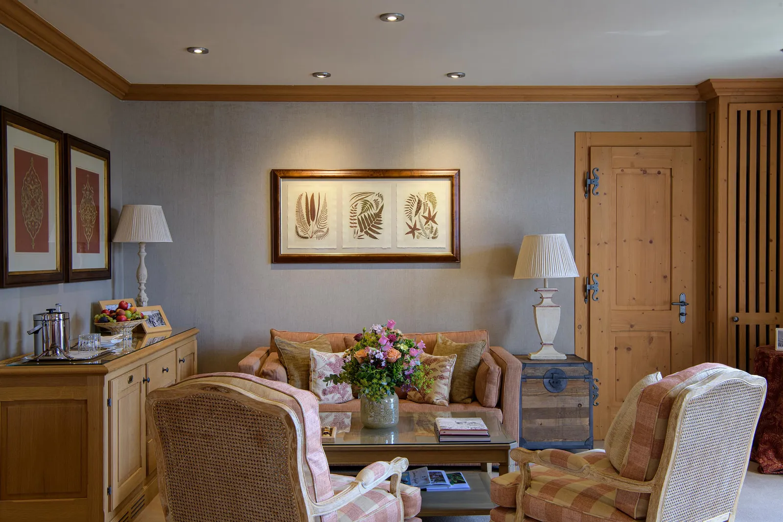 The sitting area of a Deluxe Junior Suite Mountain View: pink-checked armchairs and a sofa round a coffee table with flowers, fern prints and pine doors