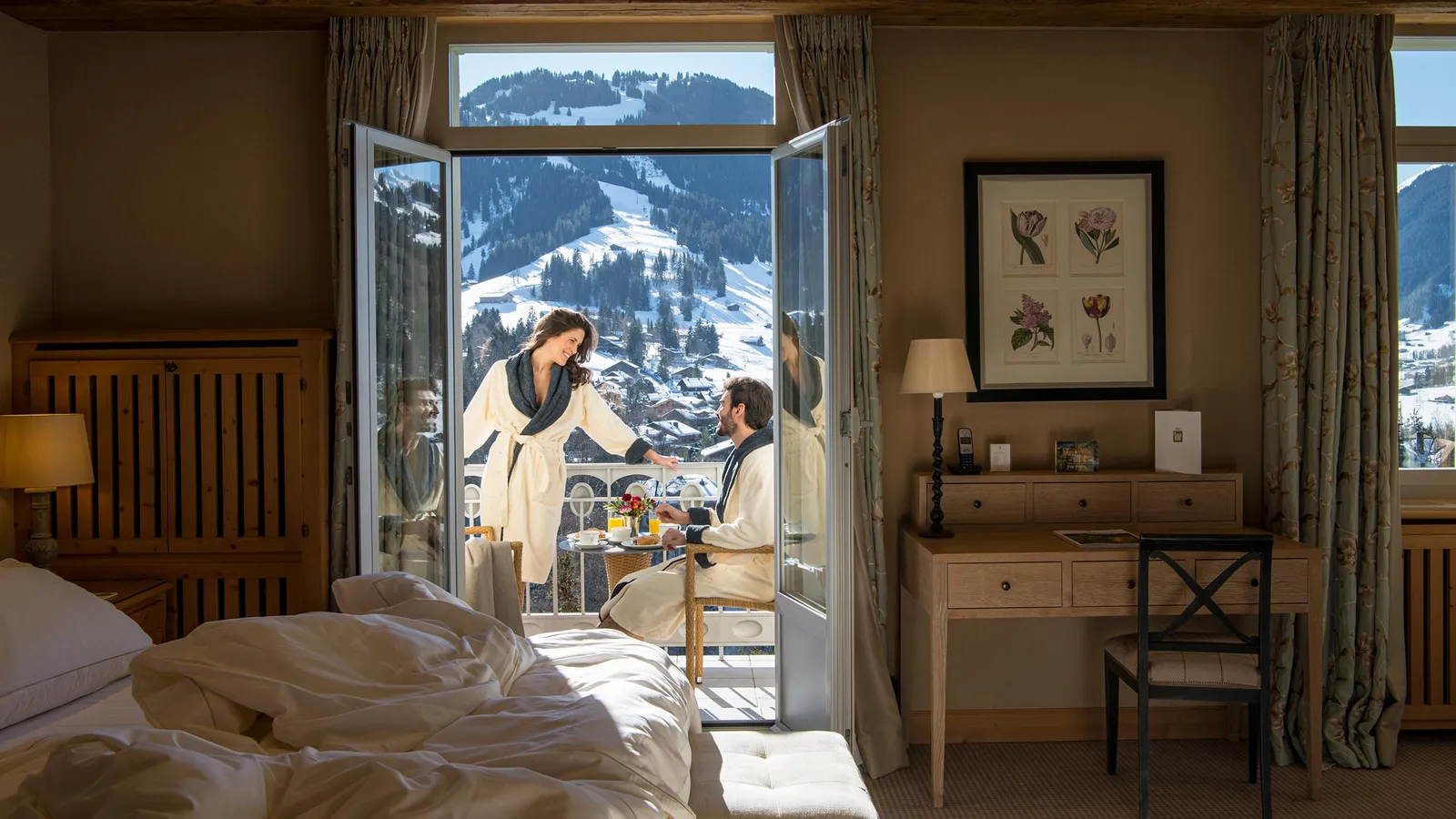 A Deluxe Room Mountain View in winter: beyond the unmade bed, a couple in bathrobes has breakfast on the balcony, with snowy slopes outside