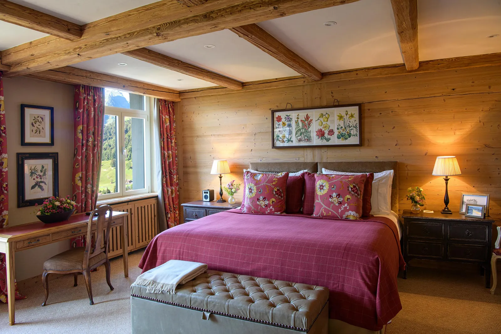 The bedroom of a Deluxe Suite Mountain View: a raspberry bedspread with embroidered cushions, pine panelling, beams, floral curtains and flower prints