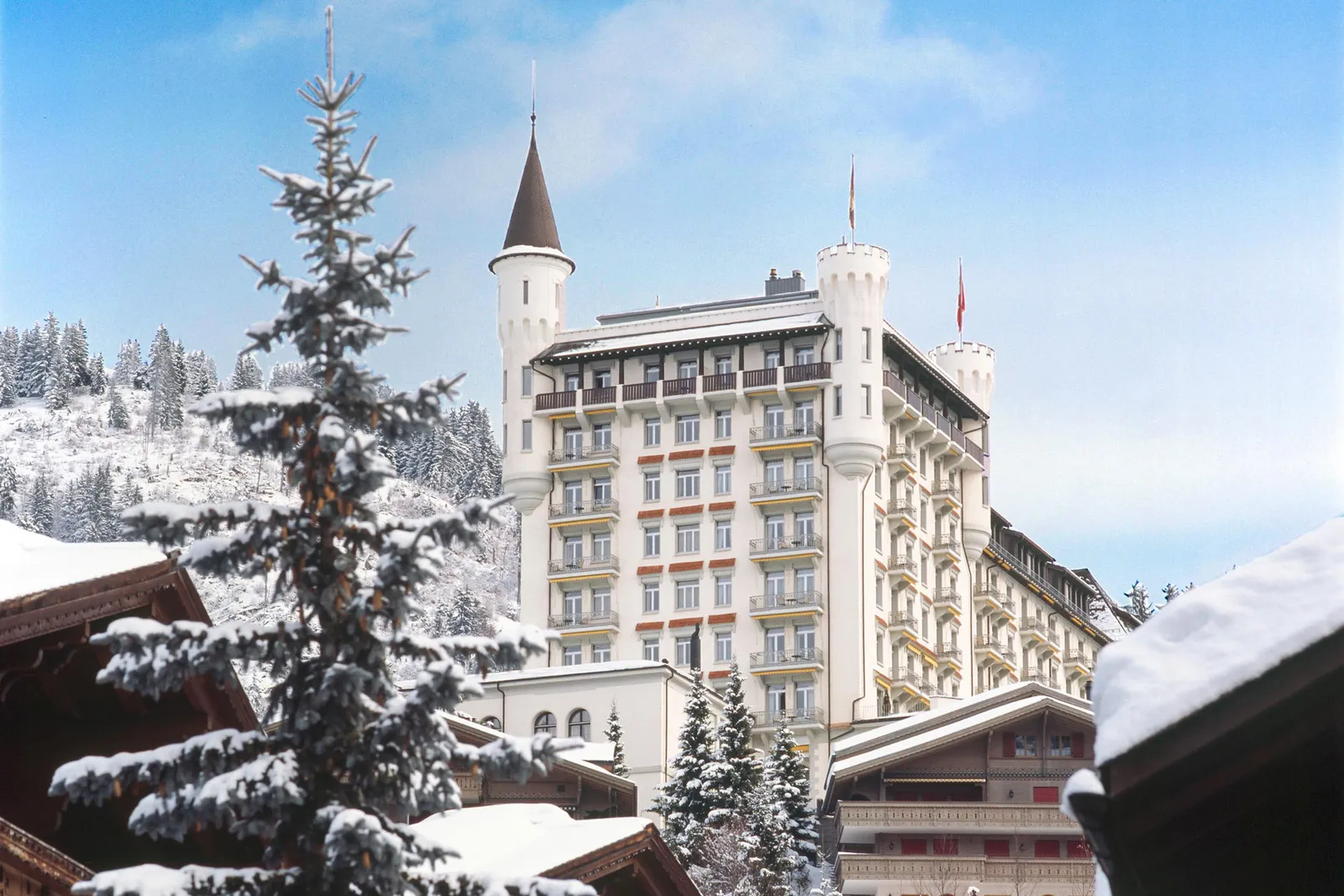 The palace and its towers in winter, seen past a snow-laden fir and the snowy roofs of chalets under a blue sky