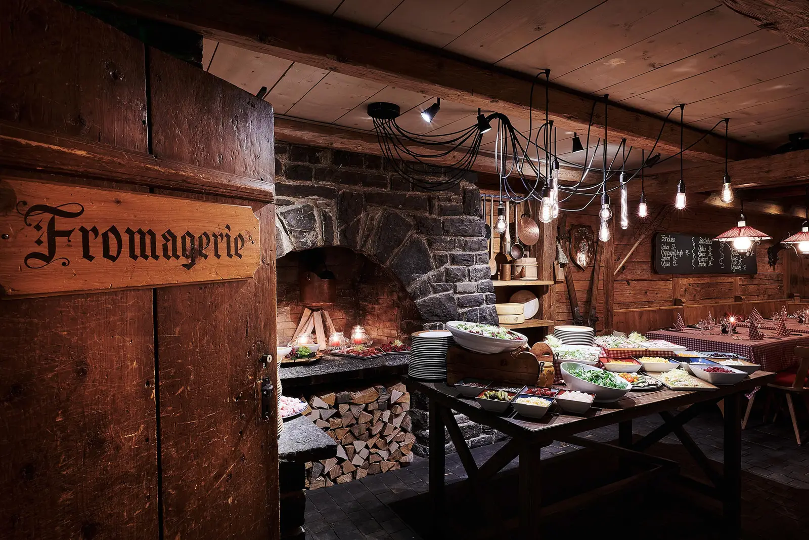 La Fromagerie: the carved sign on the wooden door, the stone vault with the raclette fire, a buffet and stacked logs