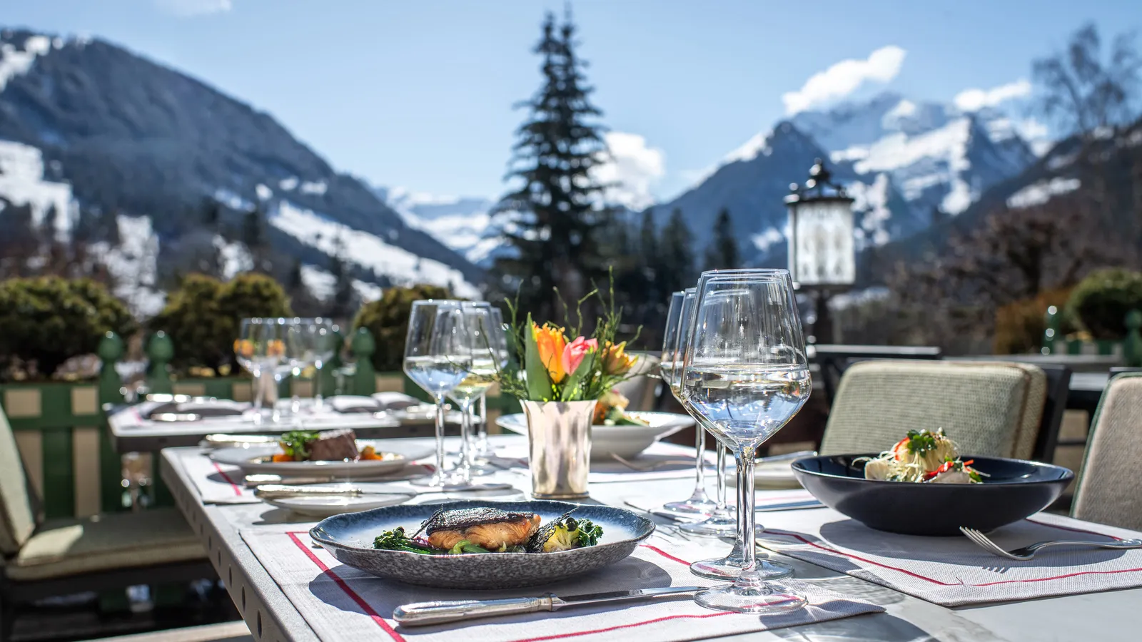 Lunch on La Terrasse at the end of winter: a laid table with plates, glasses and tulips and, beyond the railing, a fir and snowy peaks