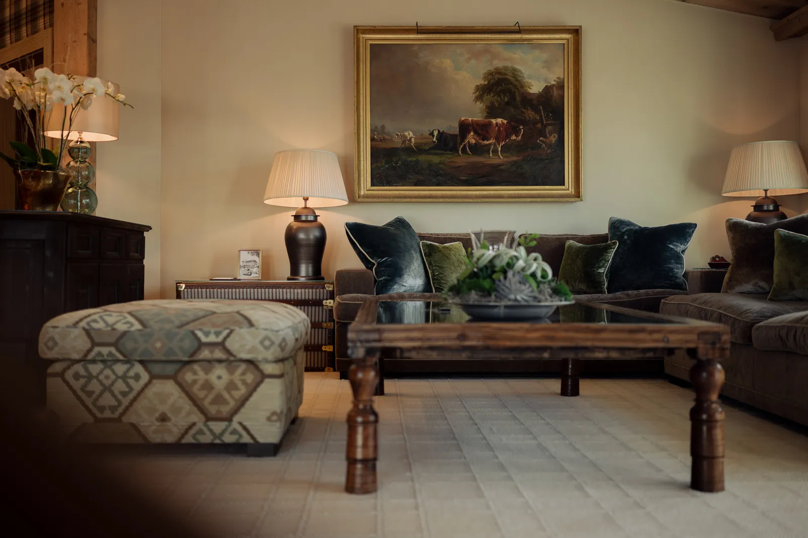 The living room of the Penthouse Suite: a large sofa with teal cushions, a patterned ottoman, a dark wooden coffee table and a painting of a cow in a landscape