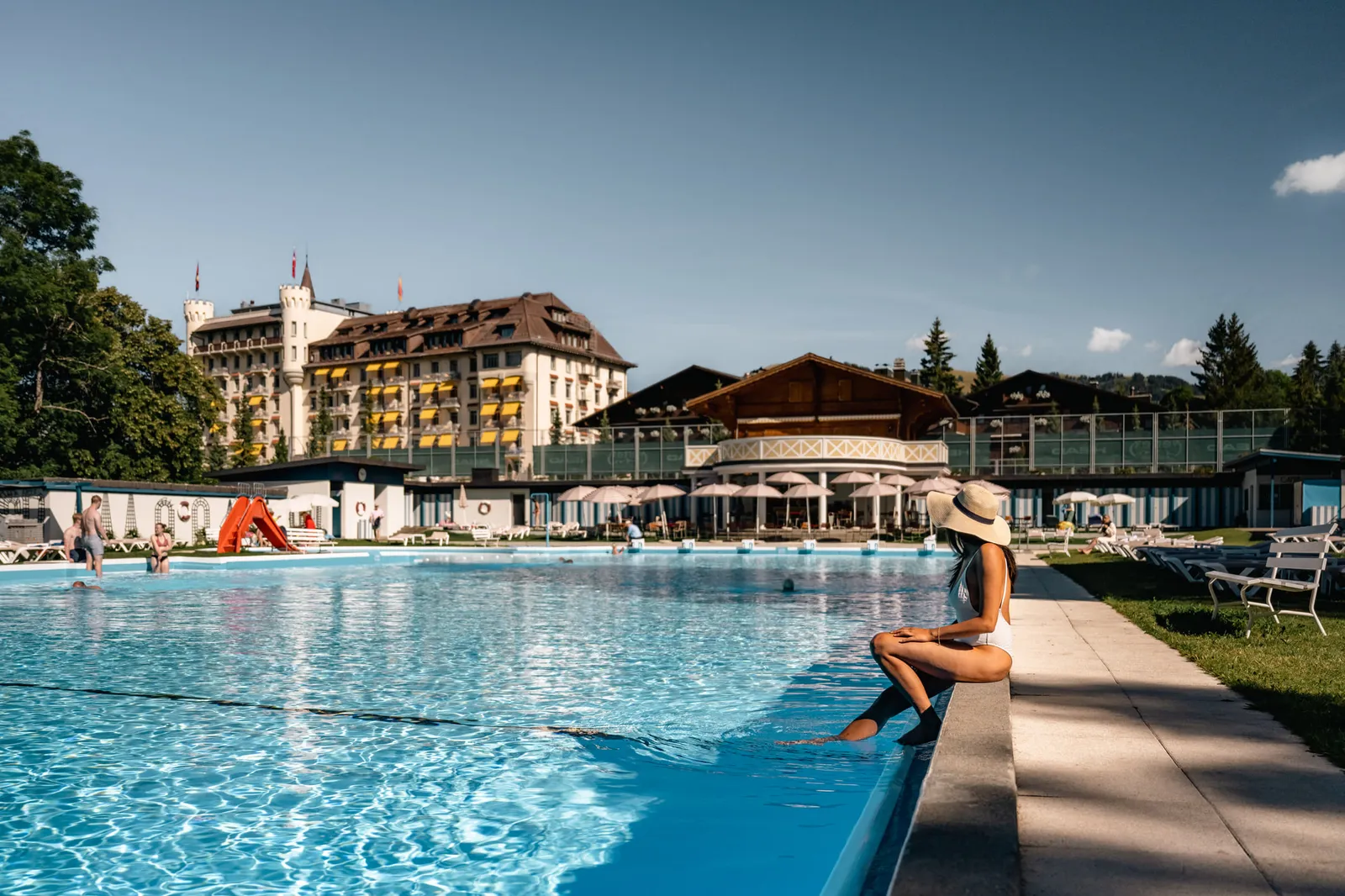 The «PISCINE» in summer: the long open-air pool, its round pavilion and the palace behind, with a woman in a sun hat sitting on the edge