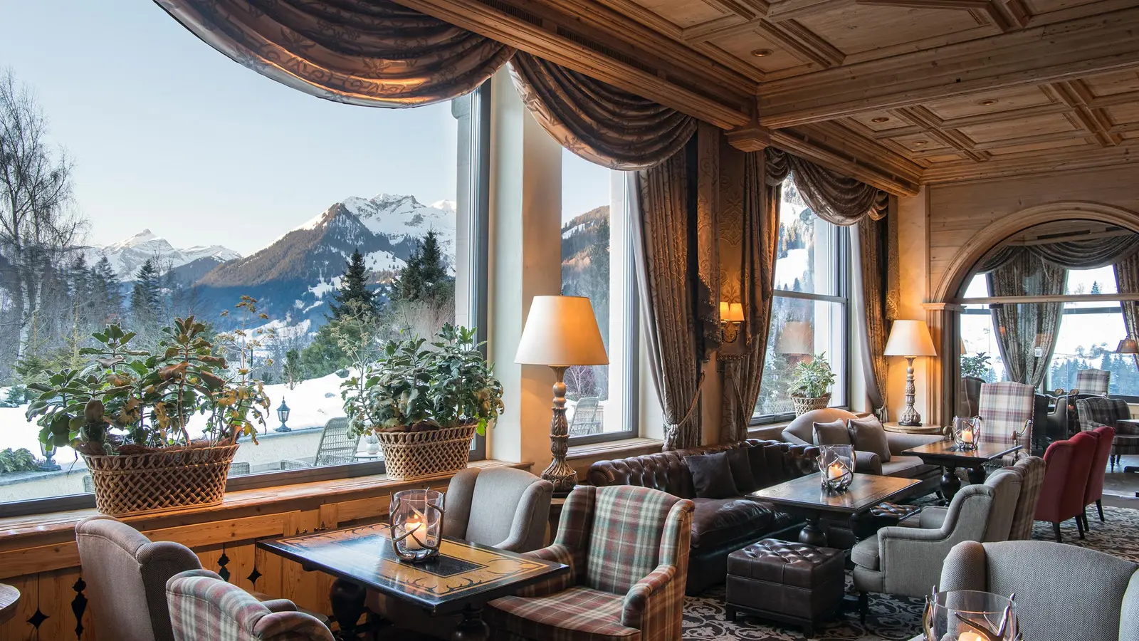 A lounge of the hotel in winter: tall windows with draped curtains onto snowy peaks, tartan and leather armchairs, lamps and candles