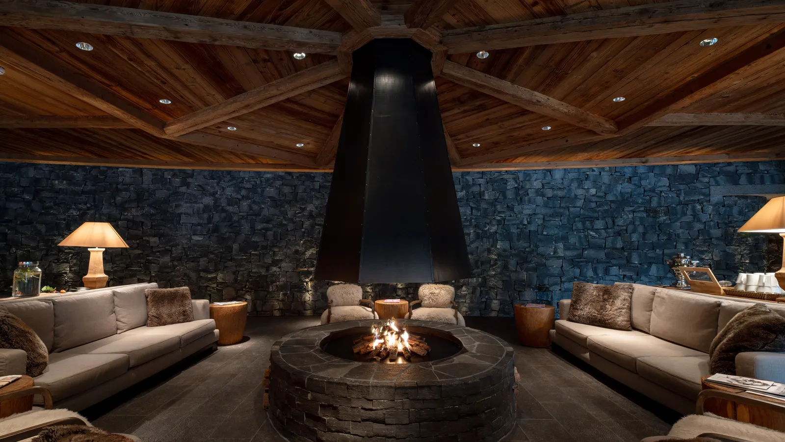 The spa lounge: a round fire pit under a black hood, sofas with fur throws, a dark stone wall and a timbered ceiling