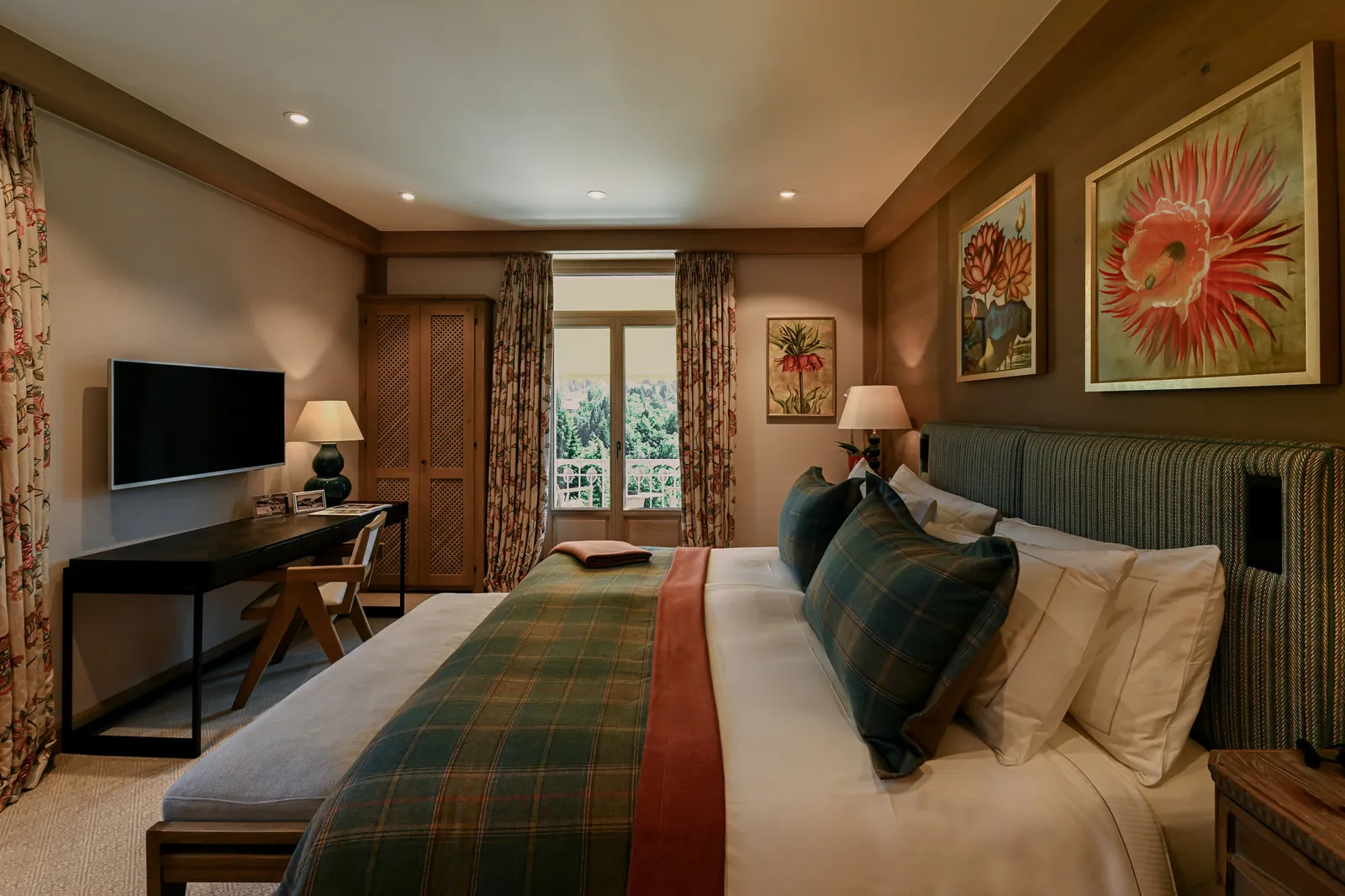 A Superior Room Mountain View: a bed with a green tartan throw and a red stripe, large flower paintings, a desk and the balcony door onto trees