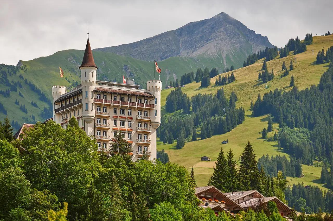 The palace in summer on its wooded hill, with turrets and flags, green pastures and a pointed peak behind and chalet roofs below