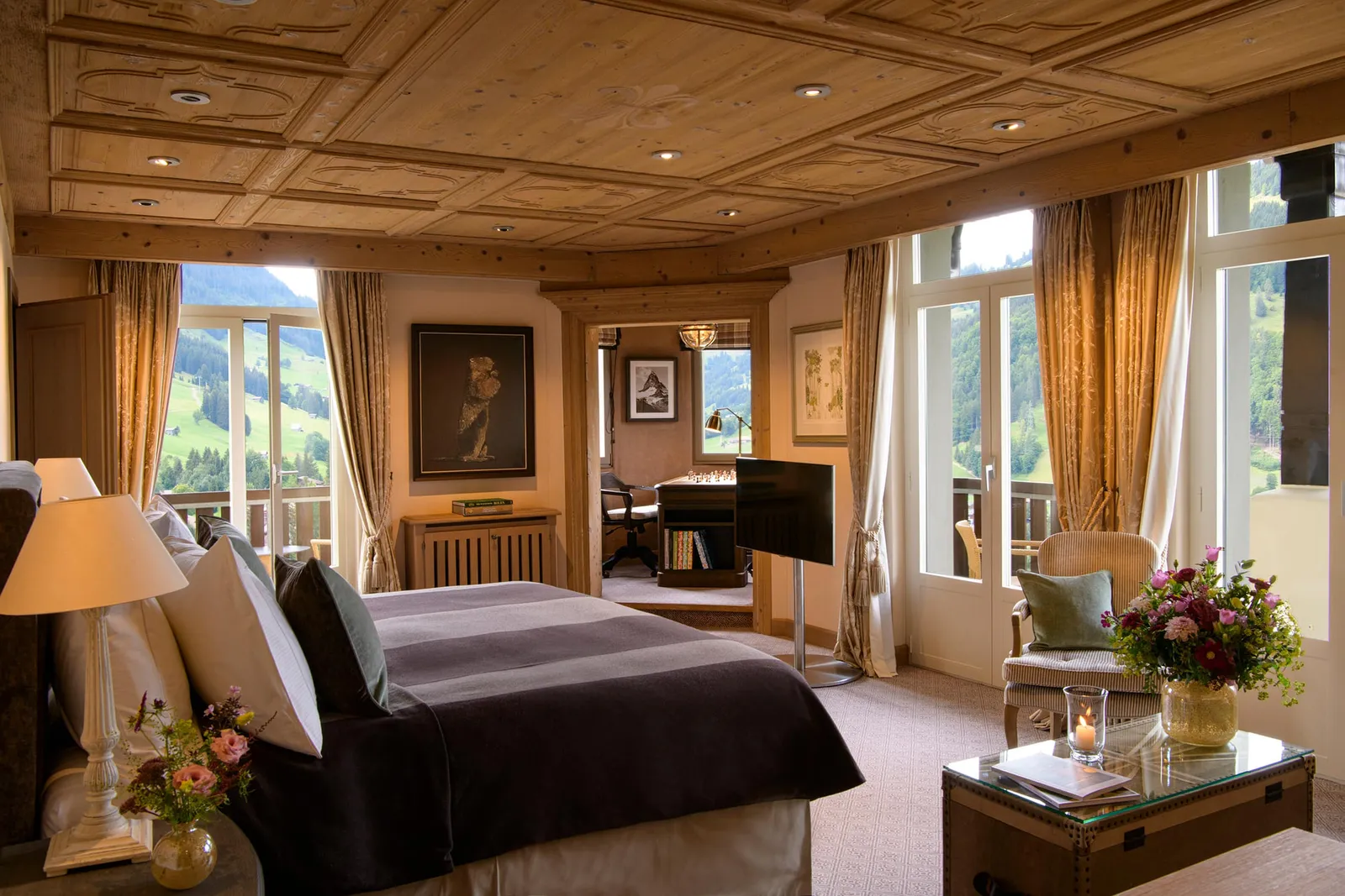 A bedroom of the Tower Two Bedroom Suite: a carved coffered pine ceiling, French windows onto balconies and green slopes and, through the door, a study